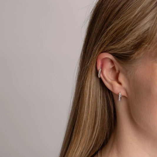 A close-up side profile of a woman's ear wearing two small silver Braid huggie hoops, one on the helix and one on the lobe, against a plain light grey background.