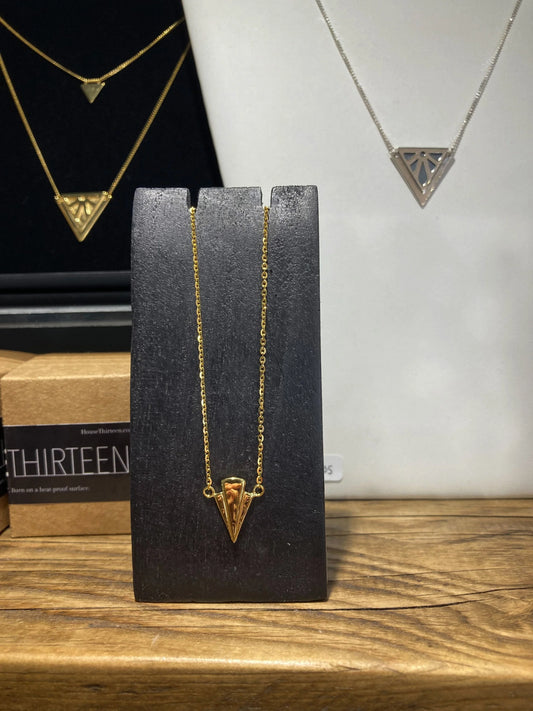 A gold necklace with a triangular fan pendant displayed on a black stand, set on a wooden surface with other jewellery pieces in the background.