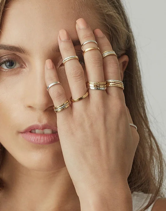 A close-up of a woman wearing multiple delicate gold rings on her fingers, with her hand covering part of her face against a soft light grey background.
