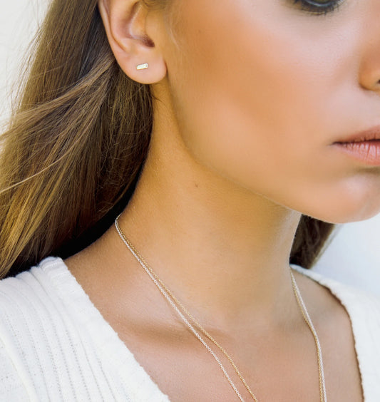 A close-up lifestyle shot of a woman wearing a delicate gold bar stud earring and a fine gold chain necklace, paired with a white ribbed top.