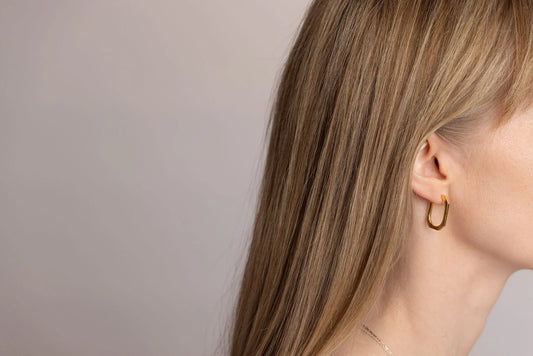A close-up side profile of a woman with long blonde hair wearing a gold Square hoop earring against a plain light grey background.