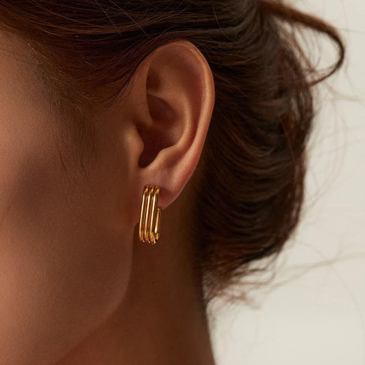 A close-up of a model's ear wearing a gold Trio hoop earring with three parallel wire bands forming a rectangular hoop, set against a soft, light neutral background.