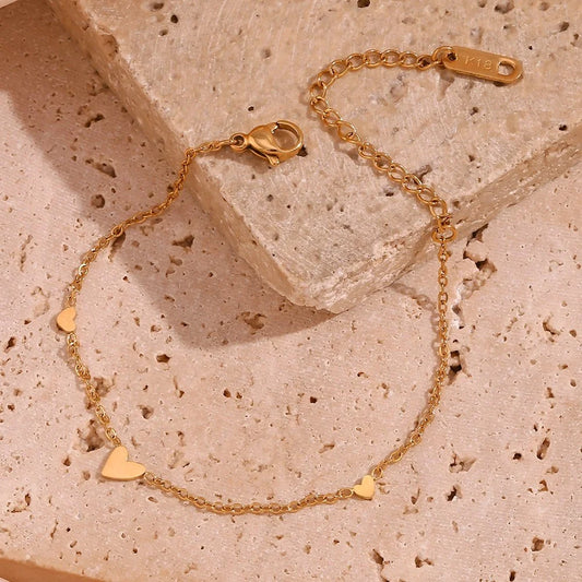A delicate gold chain bracelet with three small heart charms, displayed on a textured beige stone surface.
