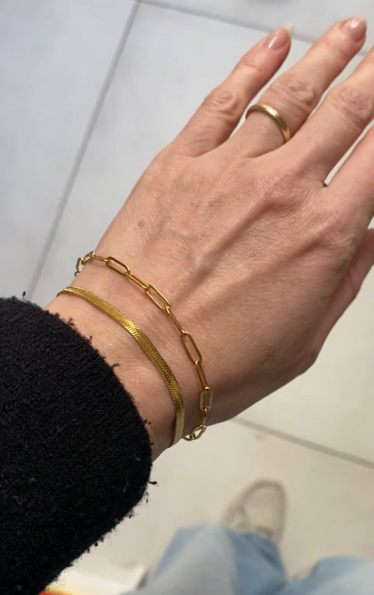 A close-up of a person's hand wearing two gold bracelets, one with a textured snake skin pattern and one with a paperclip chain link design, along with a gold ring on the finger.