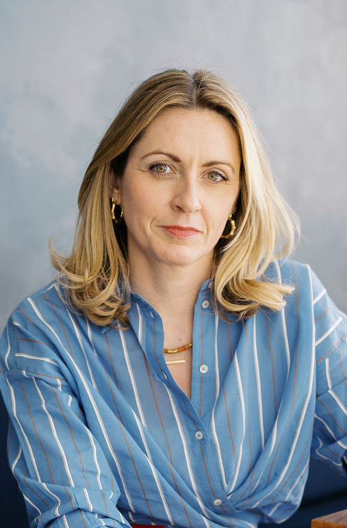 A woman with blonde hair wears a light blue and white striped button-down shirt with a yellow jade gemstone necklace and foundation bar necklace in gold and some large hoop earrings. She is posed in a modern interior setting with a textured gray wall behind her.
