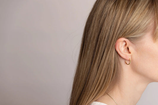 A close-up side profile of a woman with long blonde hair wearing a delicate gold Twist hoop earring, set against a soft neutral grey background.