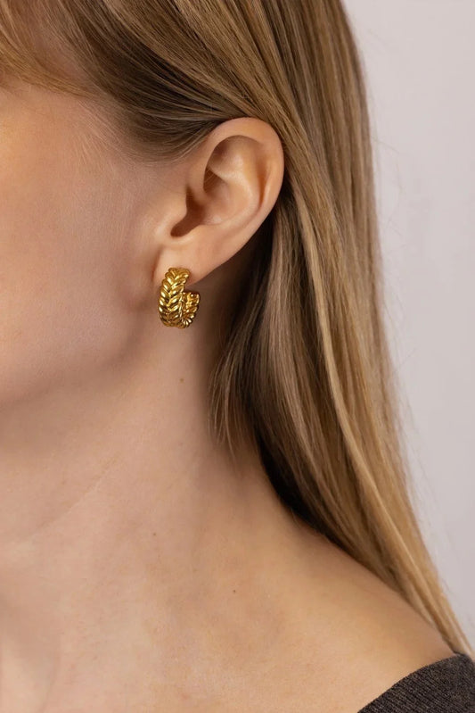 A close-up of a model's ear wearing a textured gold hoop earring against a light neutral background.