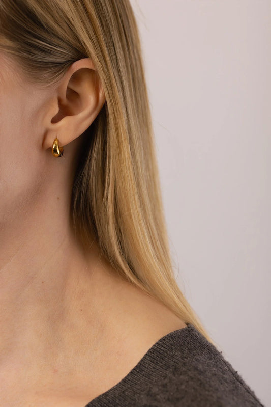 A close-up of a woman's ear wearing a small gold Cruve huggie earring against a plain light background.