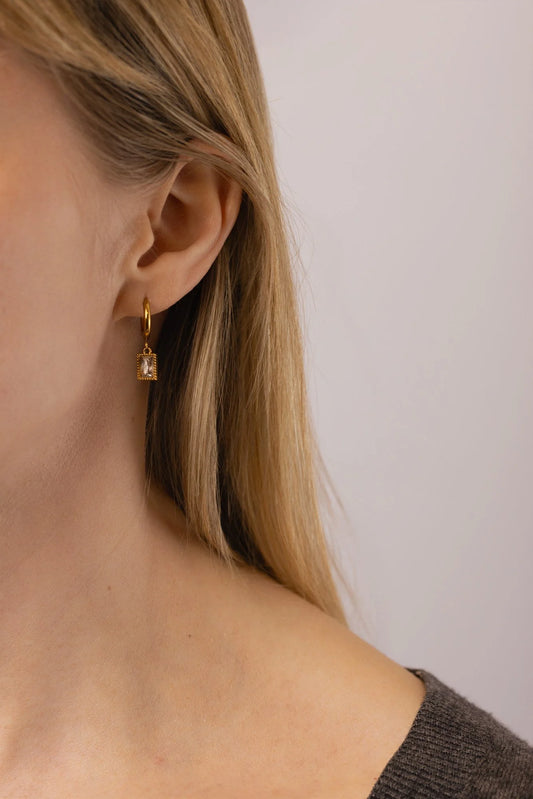 A close-up of a model's ear wearing a delicate gold Glister huggie hoop earring with a small rectangular clear cubic zirconia .