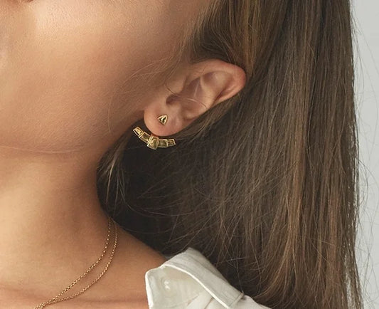 A close-up of a model's ear wearing a gold geometric fan ear jacket and a delicate gold chain necklace, with her brown hair and white shirt collar visible against a light grey background.