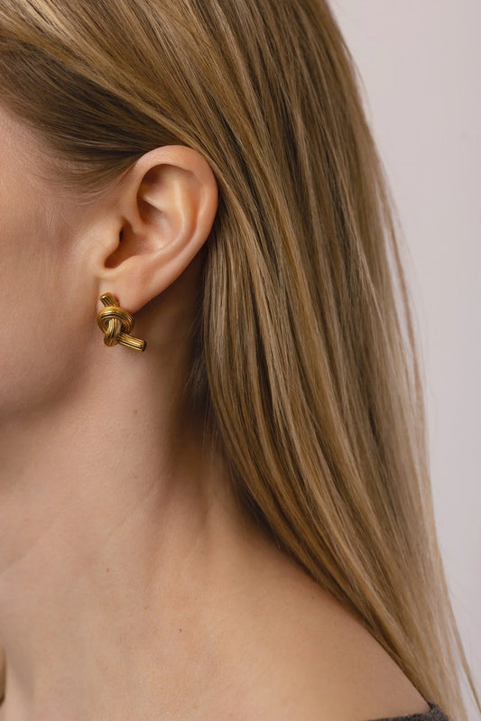 A close-up side profile of a woman with long blonde hair wearing a small gold knot-shaped stud earring against a light neutral background.