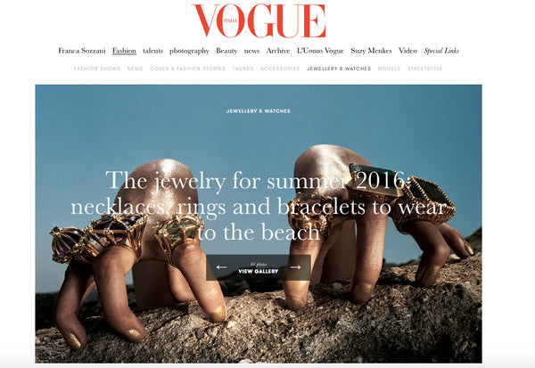 A Vogue magazine webpage screenshot featuring a jewelry cover image with multiple hands wearing ornate gold necklaces, rings, and bracelets against a blue sky and rocky beach background. The red Vogue logo is visible at the top.