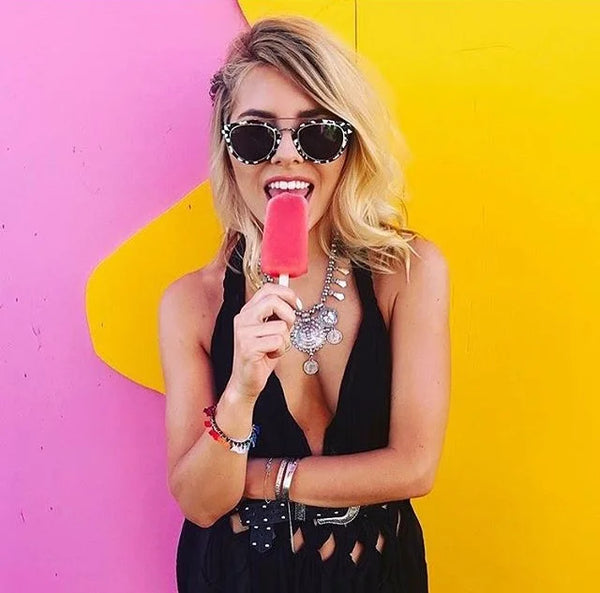 A woman, Molly King, with blonde hair wearing black sunglasses with rhinestone trim, a black cutout dress, and silver statement jewellery holds a pink popsicle in front of a bright pink and yellow painted wall. She wears House Thirteen bangles