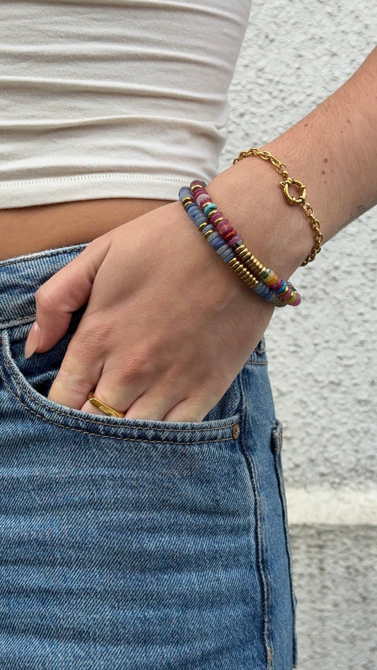 A close-up of a person's hand in a denim pocket, wearing a stack of beaded bracelets including a gold chain bracelet, a blue azurite gemstone bracelet and mixed gemstone bracelet.