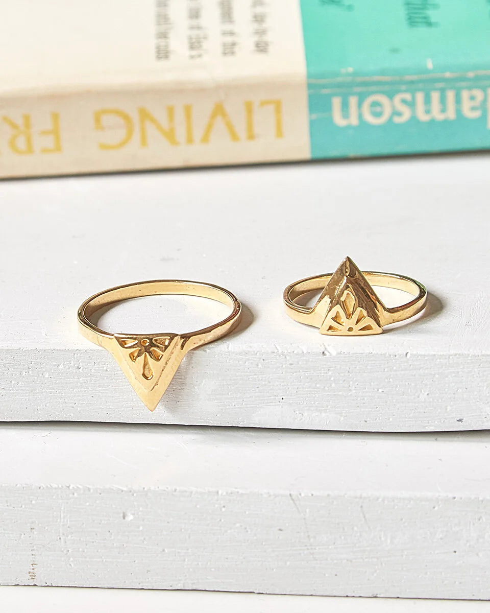 Two gold Rise rings with triangular geometric designs, displayed on a white concrete surface with a vintage book in the background.