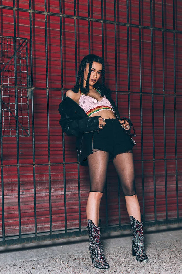 A woman posing in front of a red metal shutter, wearing a pink and green striped crop top, a black leather jacket, black denim shorts, sheer black tights, and black-and-red patterned cowboy boots.