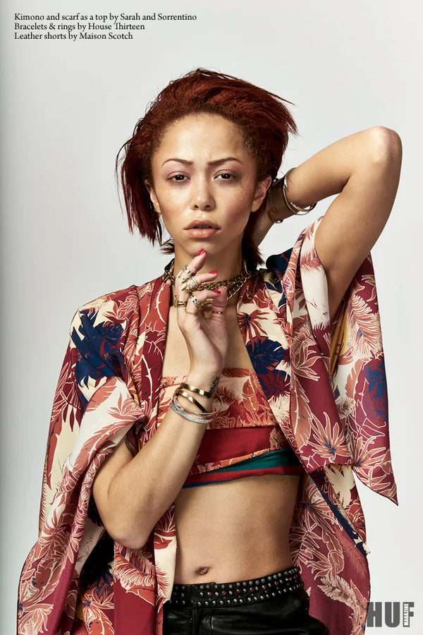 A fashion portrait of a model with short red hair wearing a floral kimono-style jacket over a red crop top and black studded shorts, with bracelets and rings, against a light gray studio background. Small text at the top left references fashion items, and a 'HUF' mark appears at the bottom right.