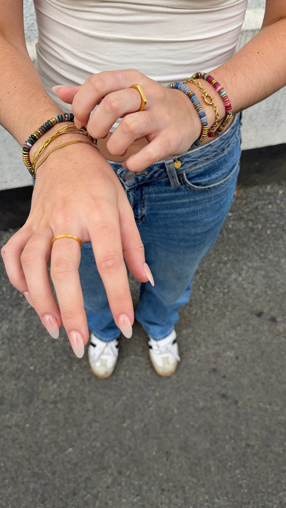A close-up of a person's hands and midsection wearing multiple gold rings and stacked gemstone bracelets, paired with a white top, blue jeans, and white sneakers on an outdoor paved surface.