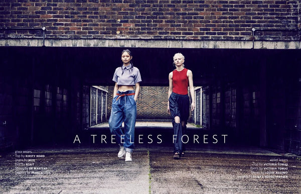 Two models walk through a dark industrial brick tunnel under a sign reading 'A TREELESS FOREST'. The model on the left wears a light purple cropped button-up shirt, loose blue jeans, and white sneakers.