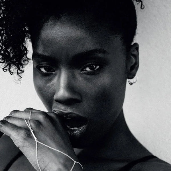 A black and white close-up portrait of a woman with her hand near her mouth, wearing delicate Drop hand chain on her fingers and wrist against a plain light background.