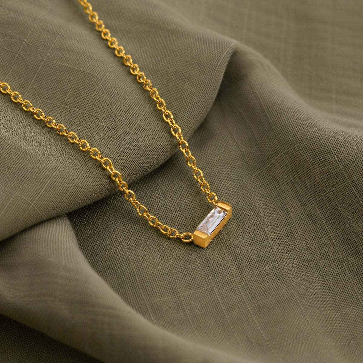 A gold chain necklace with a small rectangular cubic zirconia pendant, displayed on draped olive green fabric.