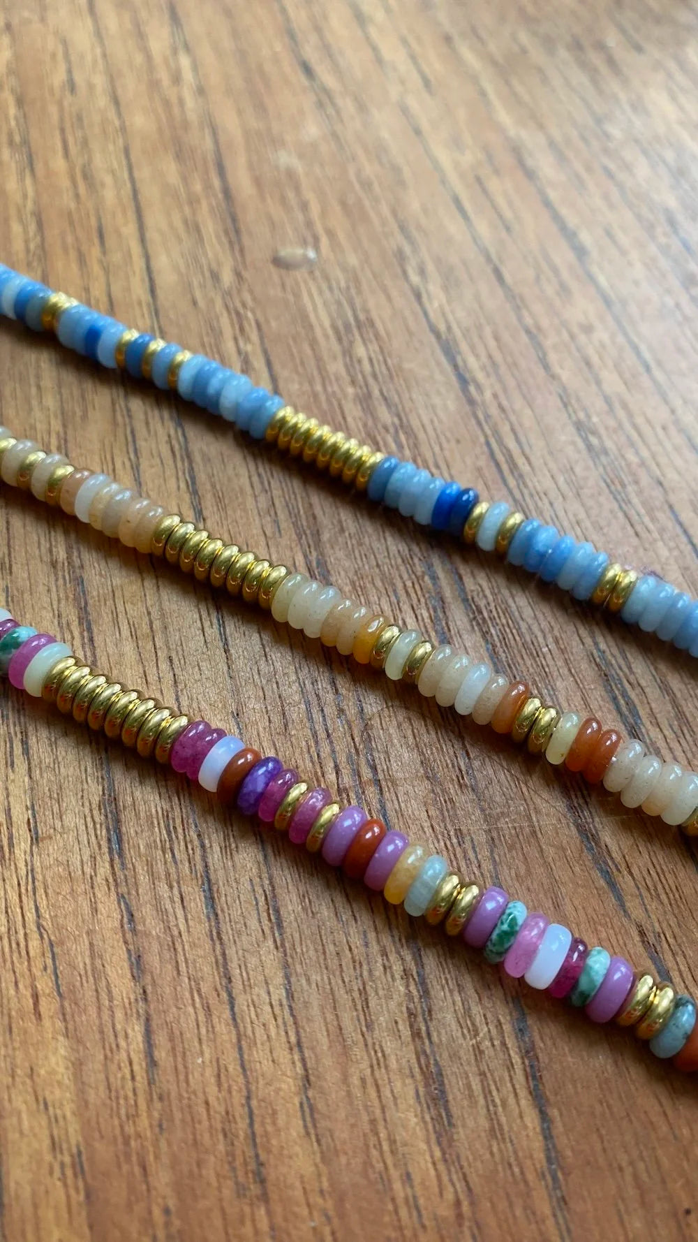 Three beaded strands of seed beads with metallic gold spacers, displayed diagonally on a brown wooden surface.