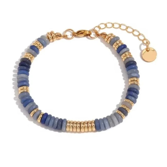 A beaded bracelet featuring alternating bands of varying tones of blue gemstones with gold beads and a gold chain extender with a round disc charm, shown on a white background.