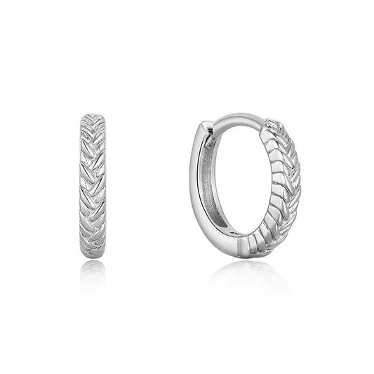 A pair of silver Braid huggie hoop earrings with a textured chevron pattern, shown from both front and side angles on a white background with soft reflections.