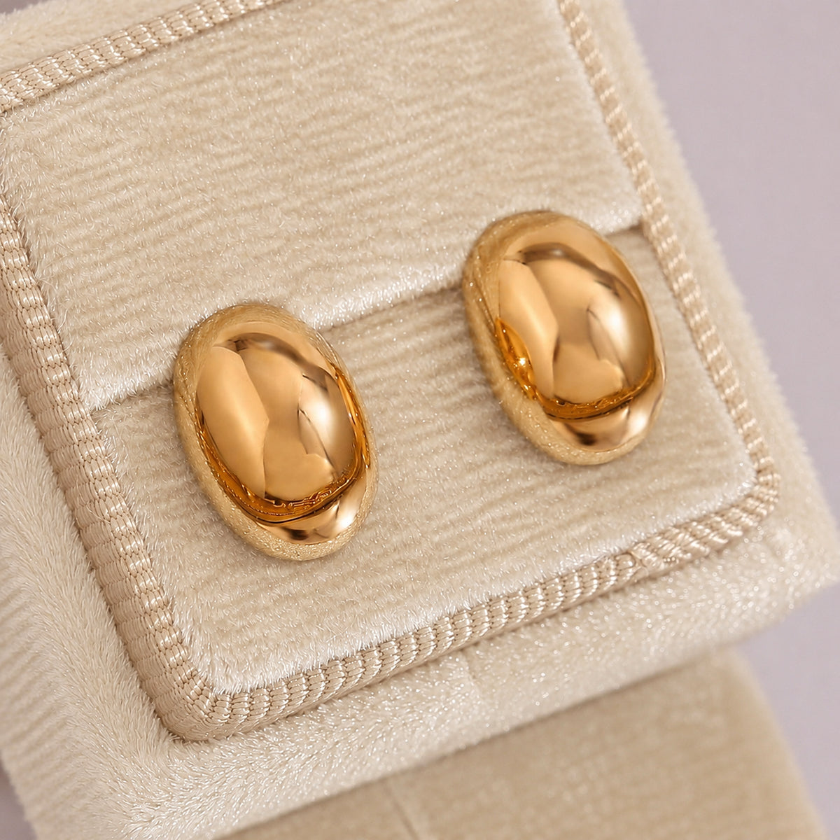 A pair of shiny gold oval stud earrings displayed on a beige velvet jewelry box.