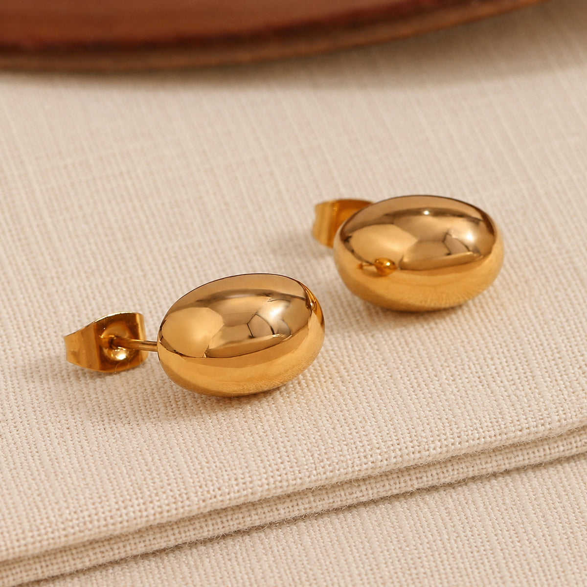 A pair of gold oval bubble stud earrings with a polished, reflective finish, resting on a textured beige fabric surface.