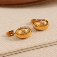 A pair of gold oval bubble stud earrings with a polished, reflective finish, resting on a textured beige fabric surface.