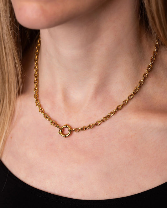 A close-up of a model's neck and chest wearing a gold chain necklace with a textured circular ring charm in the center.