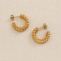 A pair of gold Chunky Braided hoop earrings with a twisted rope texture, displayed on a neutral beige textured fabric background.
