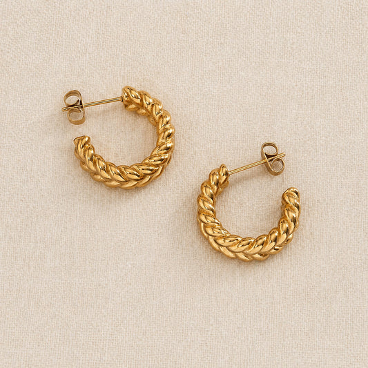 A pair of gold Chunky Braided hoop earrings with a twisted rope texture, displayed on a neutral beige textured fabric background.
