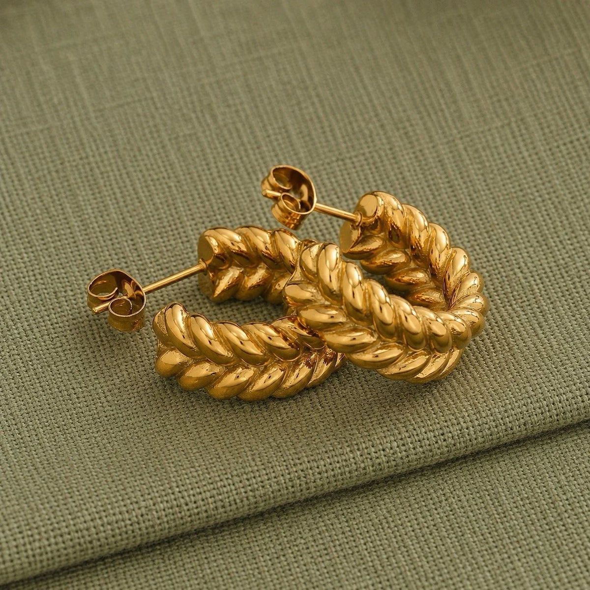 A pair of gold-toned braided hoop earrings with post-back closures, displayed on a textured olive green fabric background.