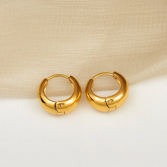 A pair of polished gold huggie hoop earrings resting on a white surface with a beige fabric backdrop.