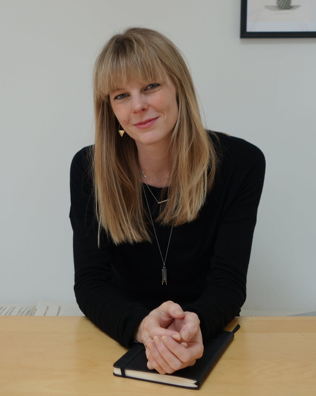 A woman (brand Founder Steph Fleming) with blonde hair sits at a light wood table in a minimalist room, wearing a black long-sleeve top. A black notebook is in front of her.