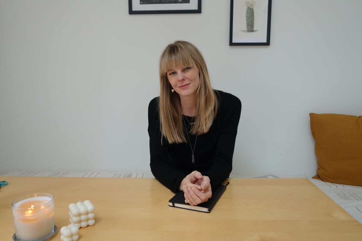 A woman with blonde hair (Founder Steph Fleming) sits at a light wood table in a minimalist room, wearing a black long-sleeve top. A black notebook is in front of her, with a lit candle and white bubble candles on the table.
