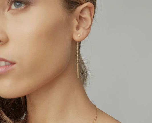 A close-up of a woman's ear wearing a delicate gold drop earring with a small round stud earring, against a plain grey background.