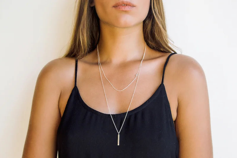 A woman wearing a layered silver necklace set, featuring a delicate chain and a longer chain with a vertical bar pendant, against a plain white background.