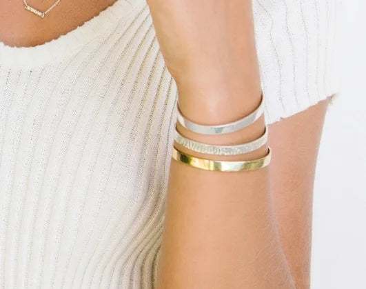 A close-up of a person's wrist wearing three stacked minimalist bangles: one polished gold, one textured silver with small stones, and one smooth silver.