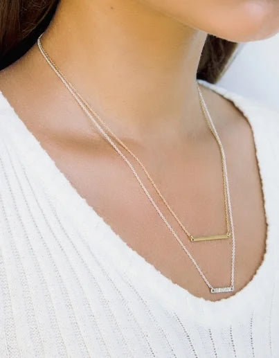 A close-up of a woman wearing a layered necklace set. The set features a delicate gold foundation bar necklace with a horizontal bar pendant and a silver bar necklace with a small rectangular textured pendant, worn over a white ribbed knit top.