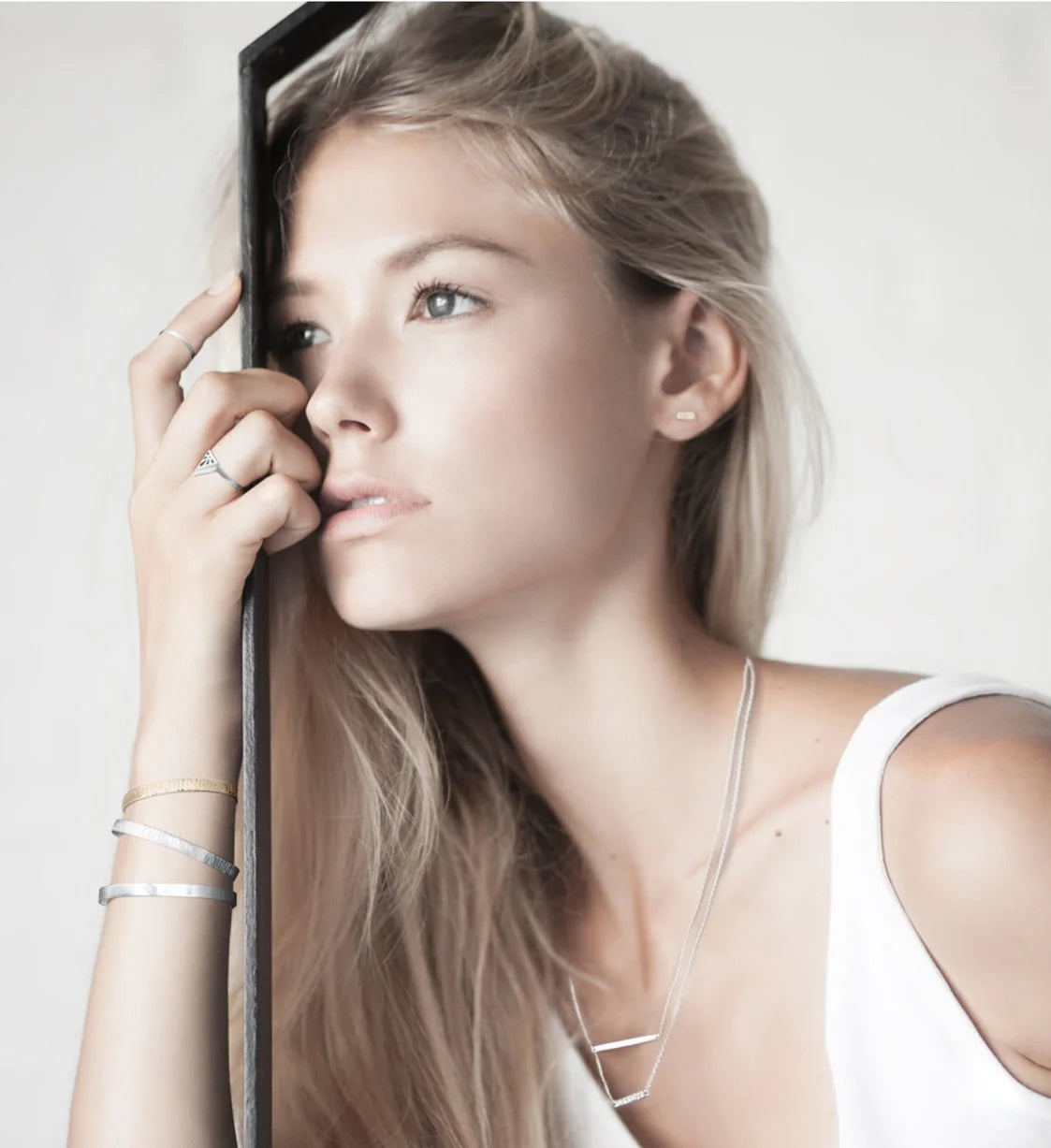 A blonde woman in a white top poses next to a black frame, showcasing delicate silver Foundation jewellery including a bar necklace, stacked bracelets, and rings.