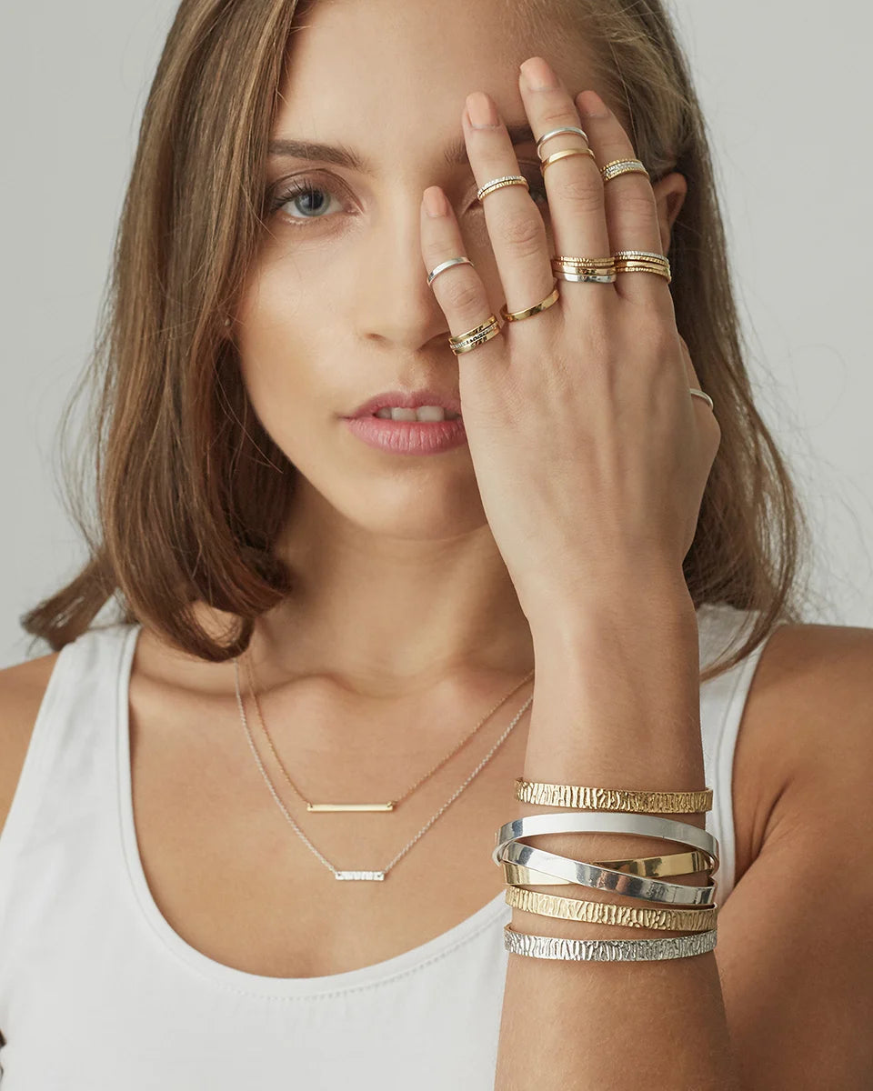 A close-up lifestyle shot of a woman wearing multiple pieces of jewelry, including gold and silver bar necklaces, stacked bangles, and several rings on her fingers, against a neutral light gray background.