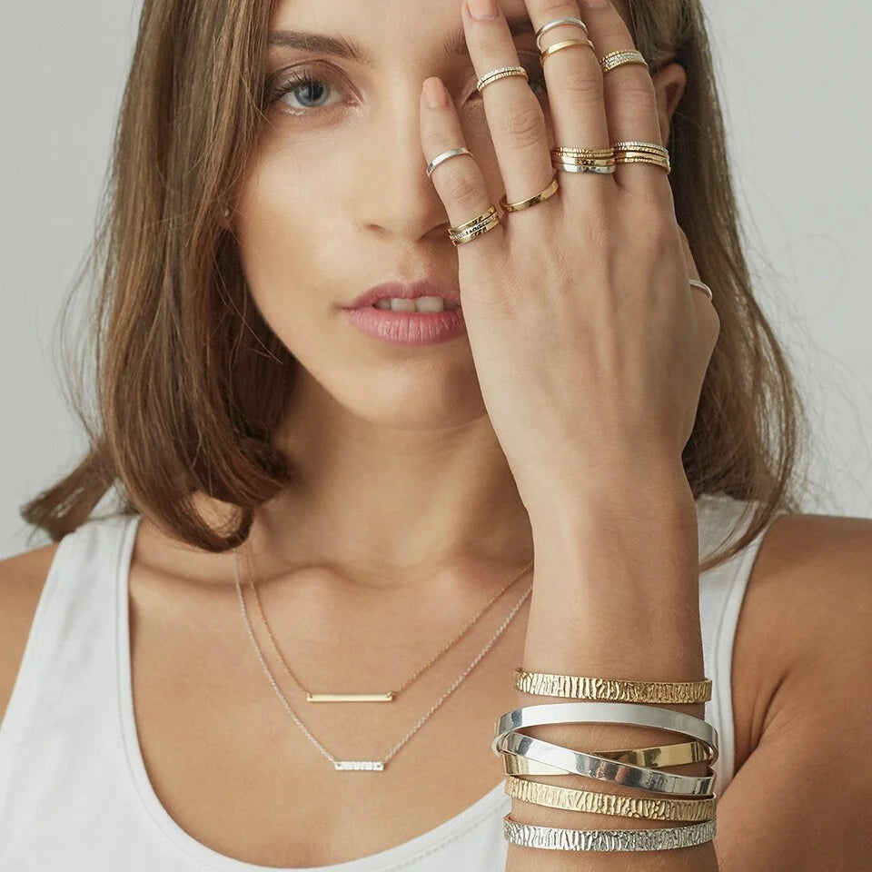 A close-up lifestyle shot of a woman wearing a stack of gold and silver bar rings on her fingers, a stack of textured gold and silver bangles on her wrist, and delicate layered gold necklaces. She is wearing a white tank top against a neutral light gray background.