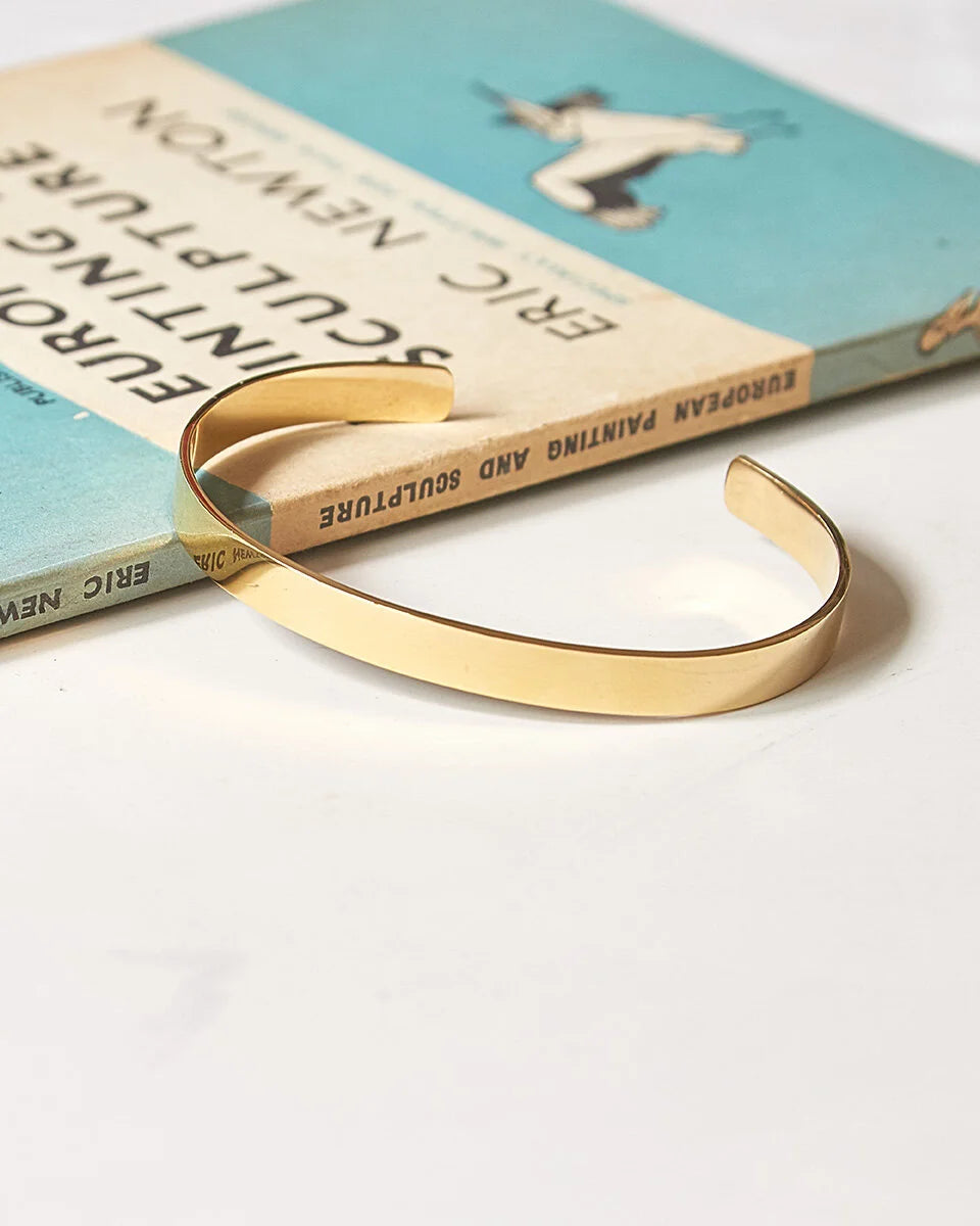 A polished gold plane bangle resting on a vintage book set against a clean white background.