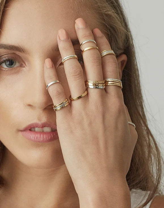A close-up of a woman wearing multiple delicate gold rings on her fingers, with her hand covering part of her face against a soft light grey background.