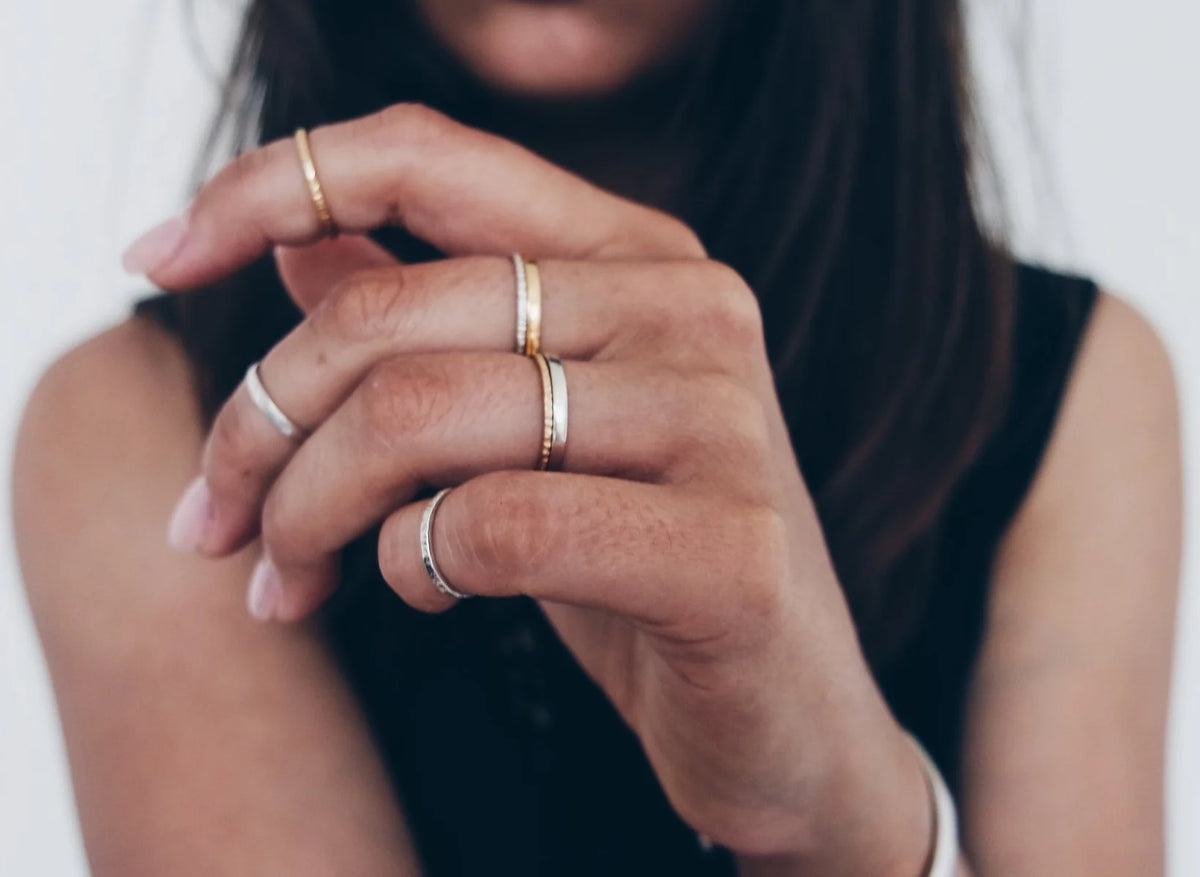 A close-up of a person's hands wearing multiple minimalist foundation rings, including two gold rings and a silver ring, against a soft light grey background.