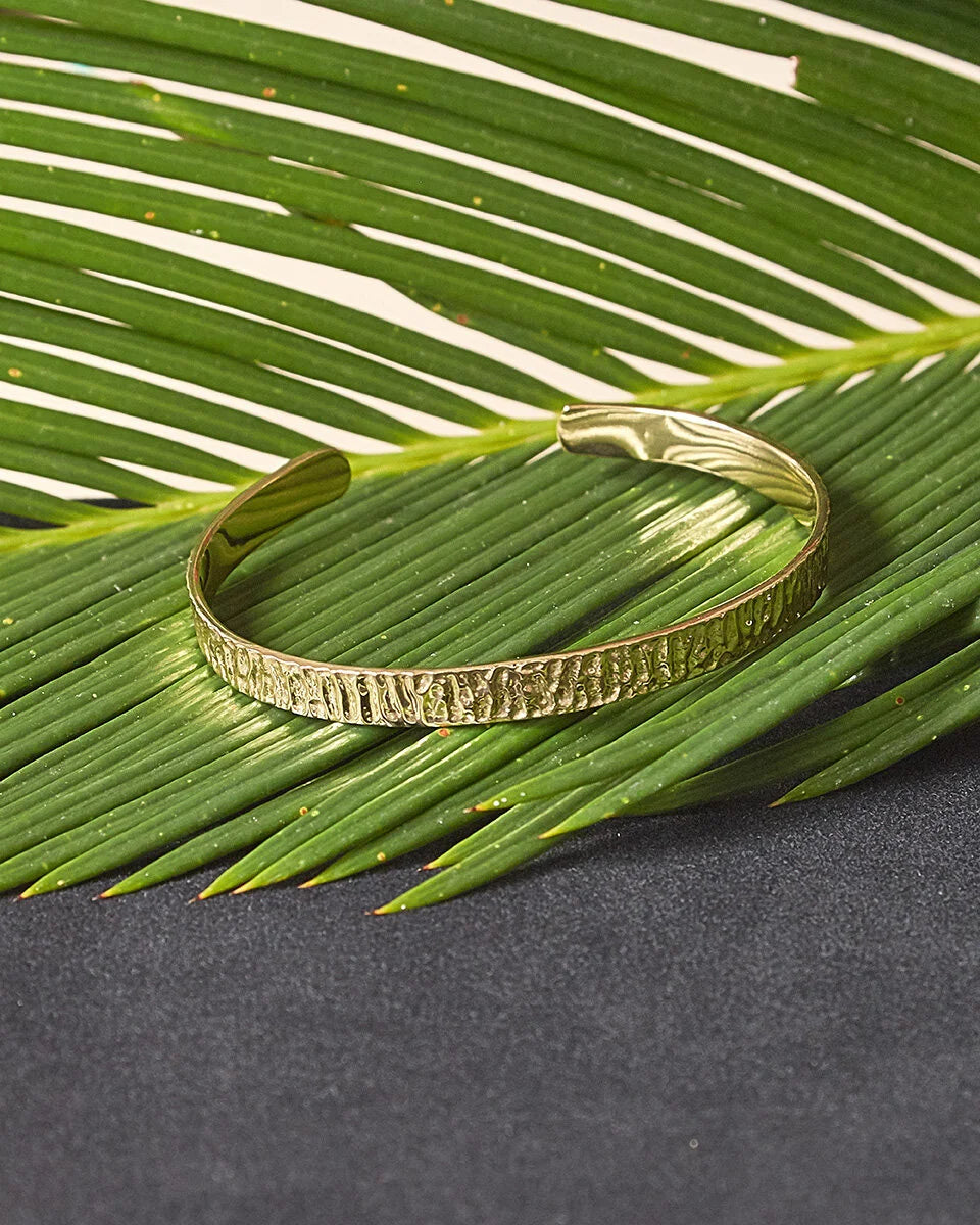 A textured gold cuff bracelet resting on a green palm leaf over a dark gray surface.
