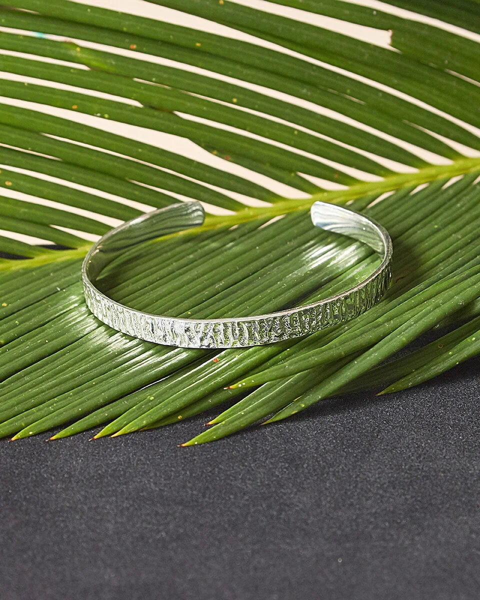 A textured silver cuff bracelet resting on a green palm leaf over a dark gray surface.